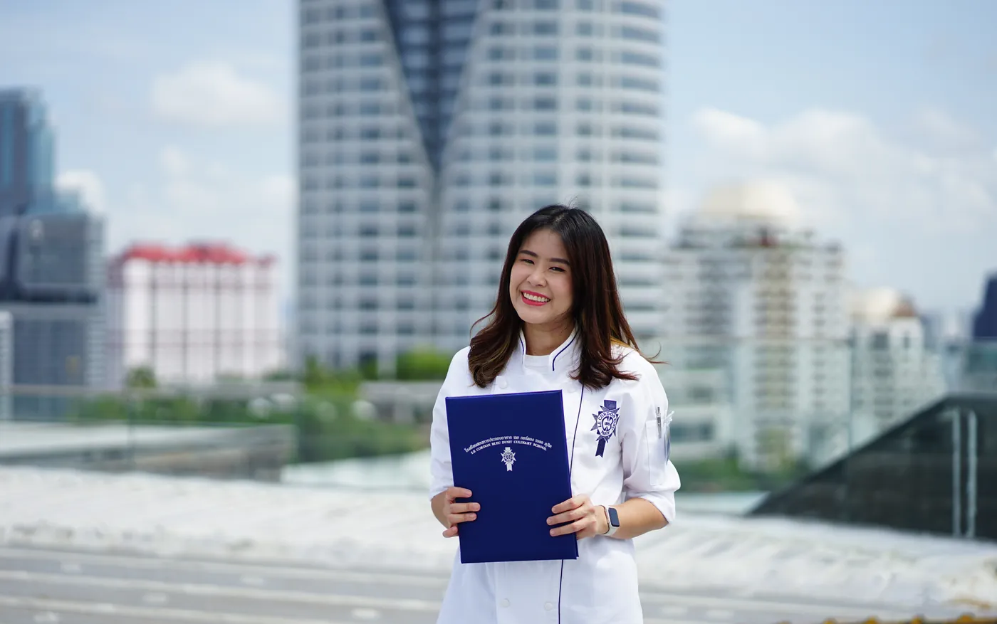 Ray's pastry chef in her chef's jacket, holding her Le Cordon Bleu Dusit diploma.
