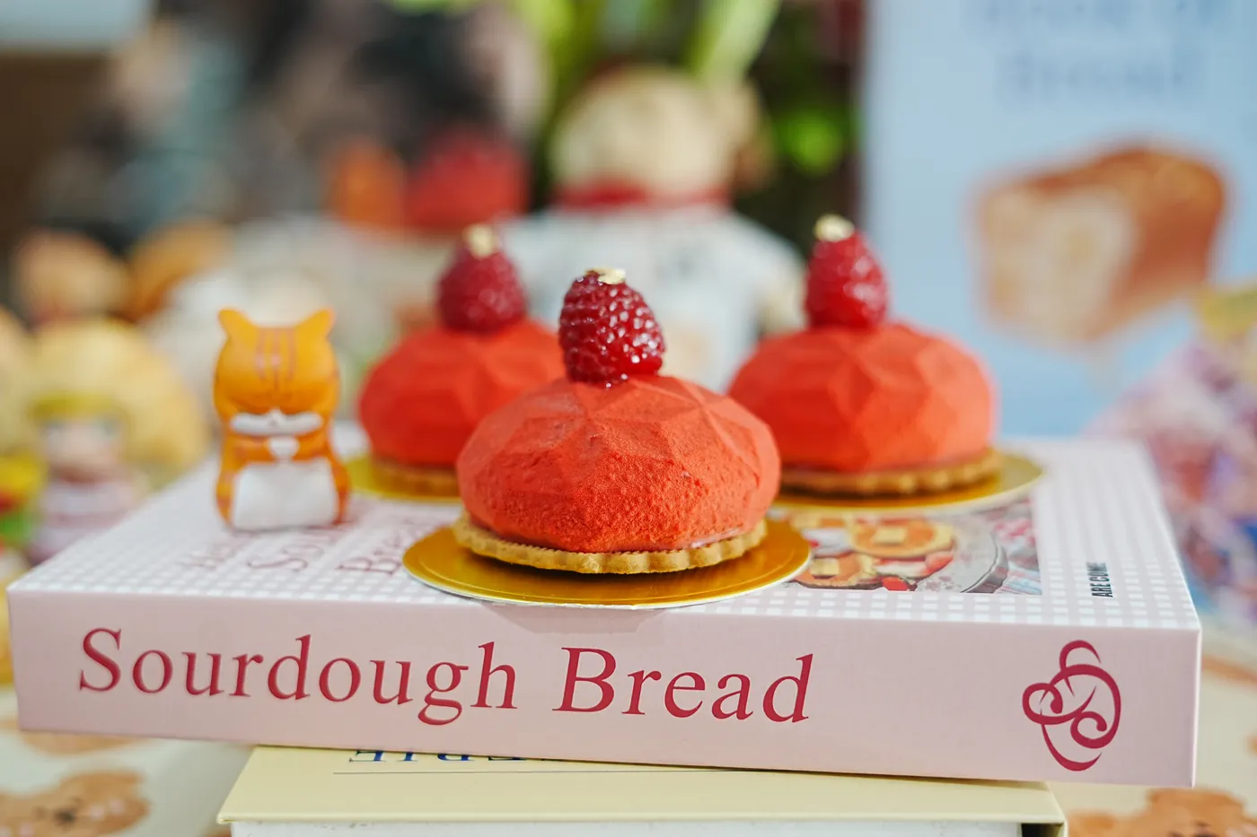 Raspberry mousse cakes on gold boards, beside a book on sourdough bread.