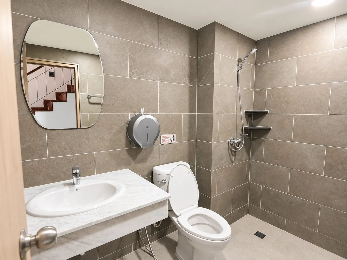 A shared bathroom: a basin under a round mirror, a toilet and a shower, all in grey tile.